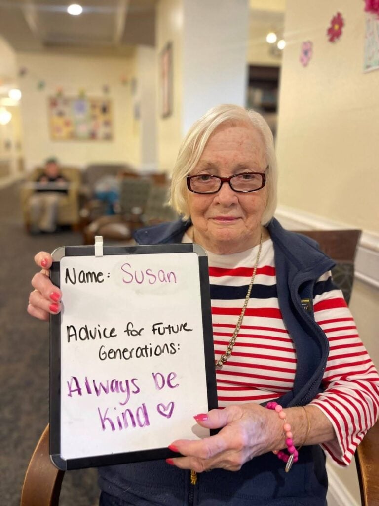 Susan sits in a chair at Standish Village and holds a whiteboard that reads "always be kind".