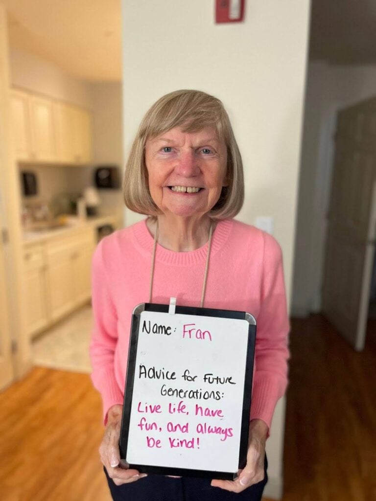 Fran smiles in her apartment and holds a whiteboard that reads "live life, have fun, and always be kind".