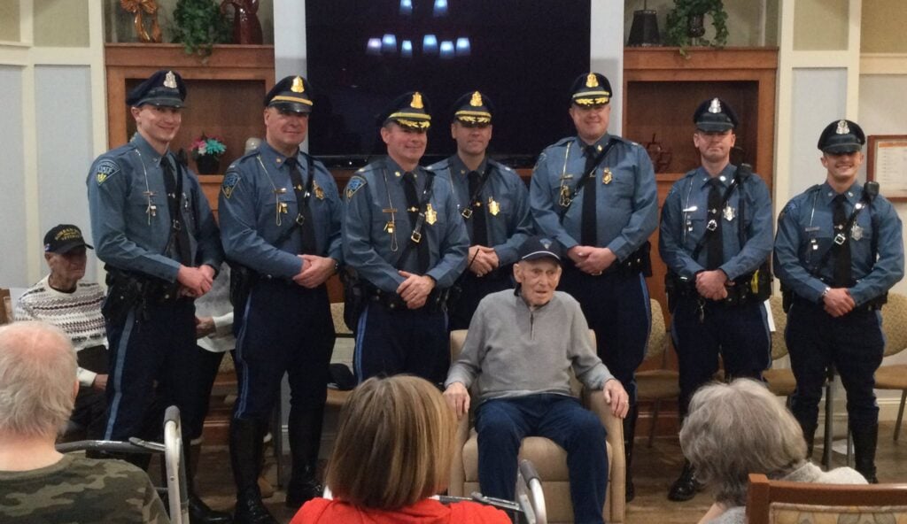 Seven Massachusetts State Police troopers in dress uniform stand behind an elderly seated resident being honored at a senior living community event, with other residents looking on.