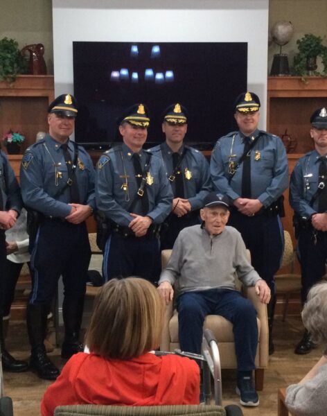 Seven Massachusetts State Police troopers in dress uniform stand behind an elderly seated resident being honored at a senior living community event, with other residents looking on.
