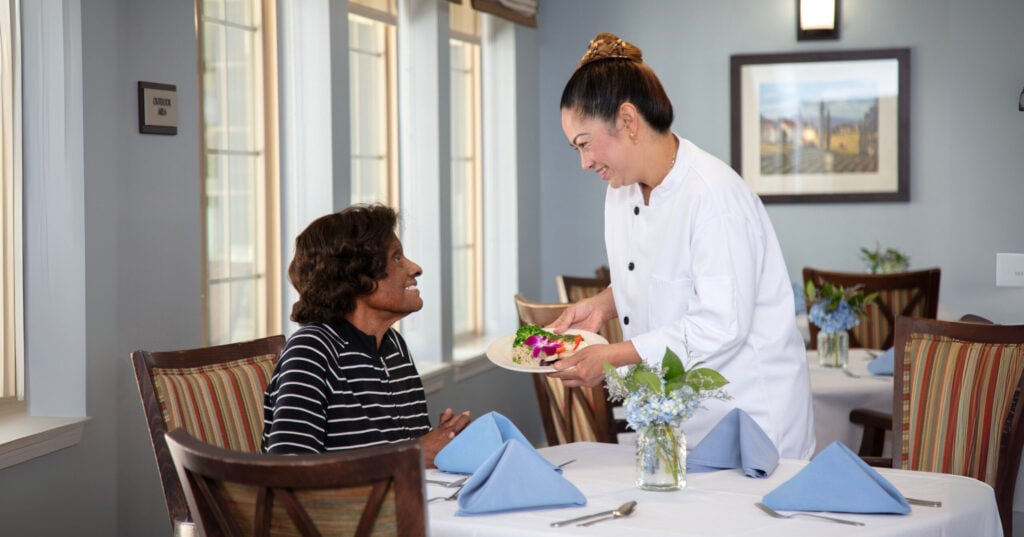 A chef in a white uniform smiles while presenting a plated meal garnished with flowers to a seated dining room guest, in an elegantly set restaurant with blue hydrangea centerpieces and large windows.