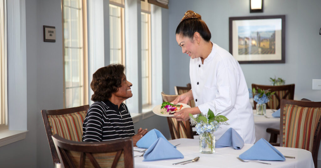 A chef in a white uniform smiles while presenting a plated meal garnished with flowers to a seated dining room guest, in an elegantly set restaurant with blue hydrangea centerpieces and large windows.