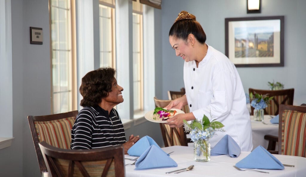 A chef in a white uniform smiles while presenting a plated meal garnished with flowers to a seated dining room guest, in an elegantly set restaurant with blue hydrangea centerpieces and large windows.