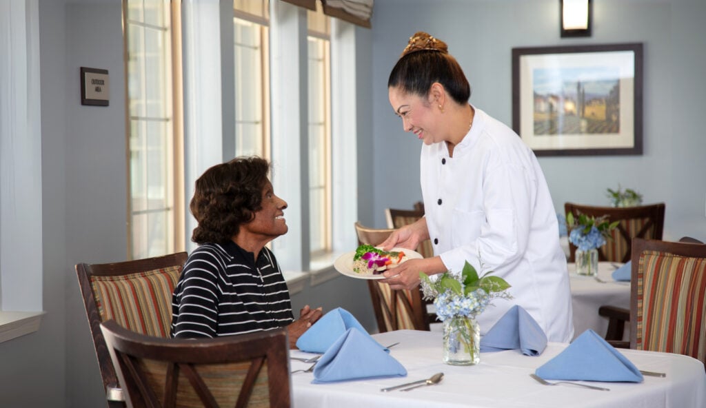 A chef in a white uniform smiles while presenting a plated meal garnished with flowers to a seated dining room guest, in an elegantly set restaurant with blue hydrangea centerpieces and large windows.