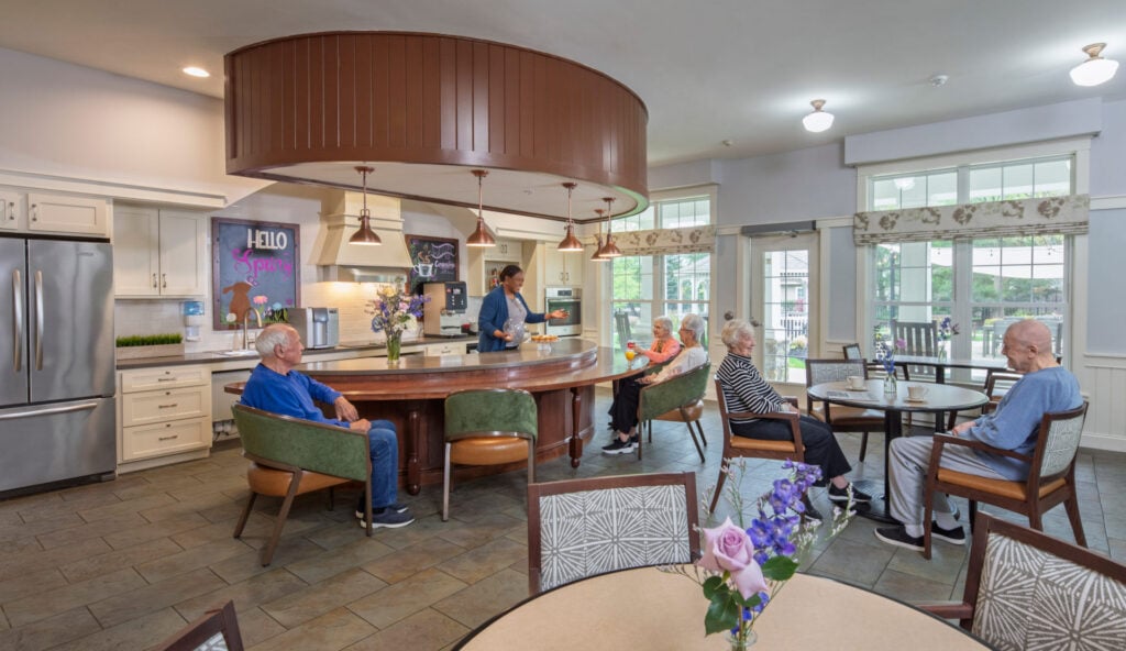 Several senior residents relax at tables and around a large curved kitchen island while a staff member serves refreshments in a bright, open community kitchen and dining area.