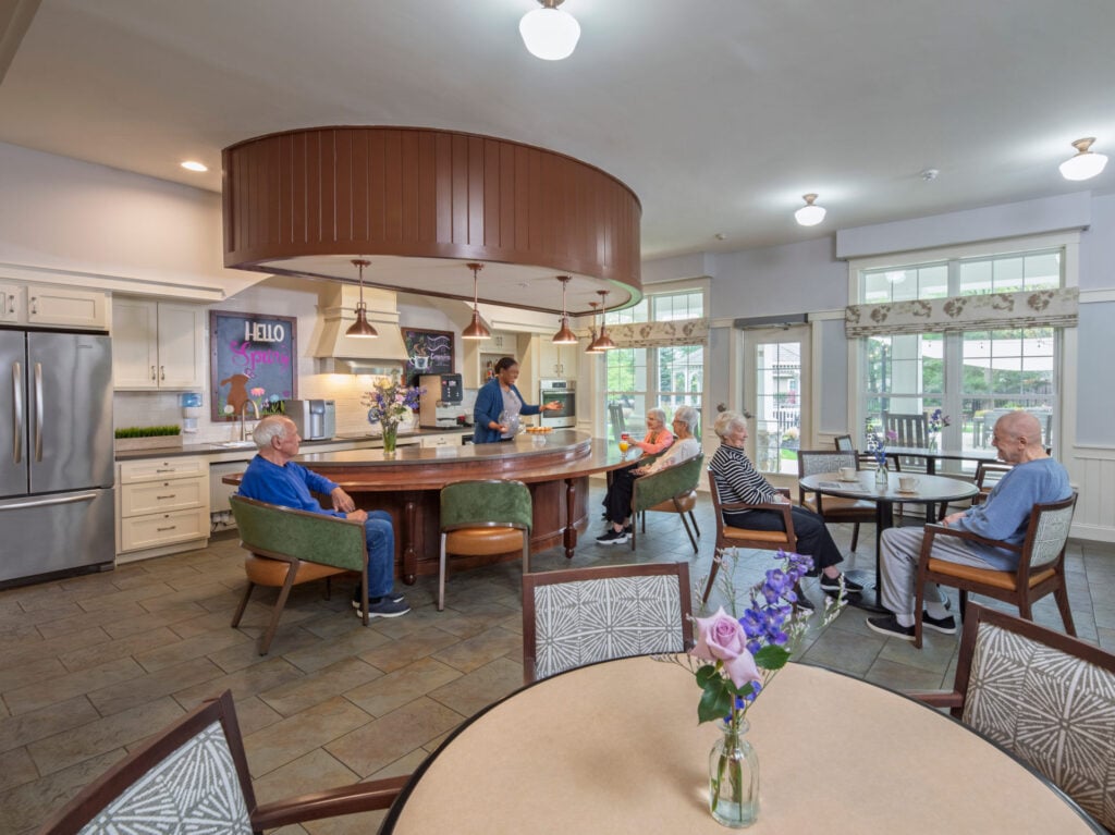 Several senior residents relax at tables and around a large curved kitchen island while a staff member serves refreshments in a bright, open community kitchen and dining area.