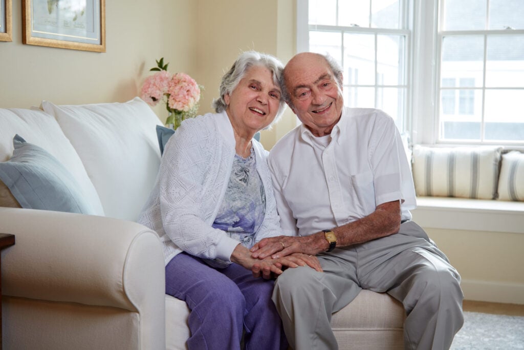 An elderly couple sits close together on a white sofa, smiling warmly and holding hands in a sunlit living room with pink hydrangeas in the background.