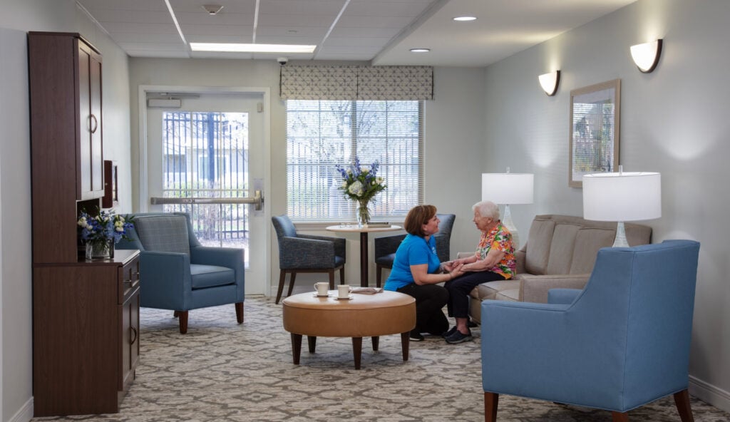 A caregiver in a blue polo shirt sits with an elderly resident on a sofa in a bright, warmly furnished common room lounge, engaged in friendly conversation.