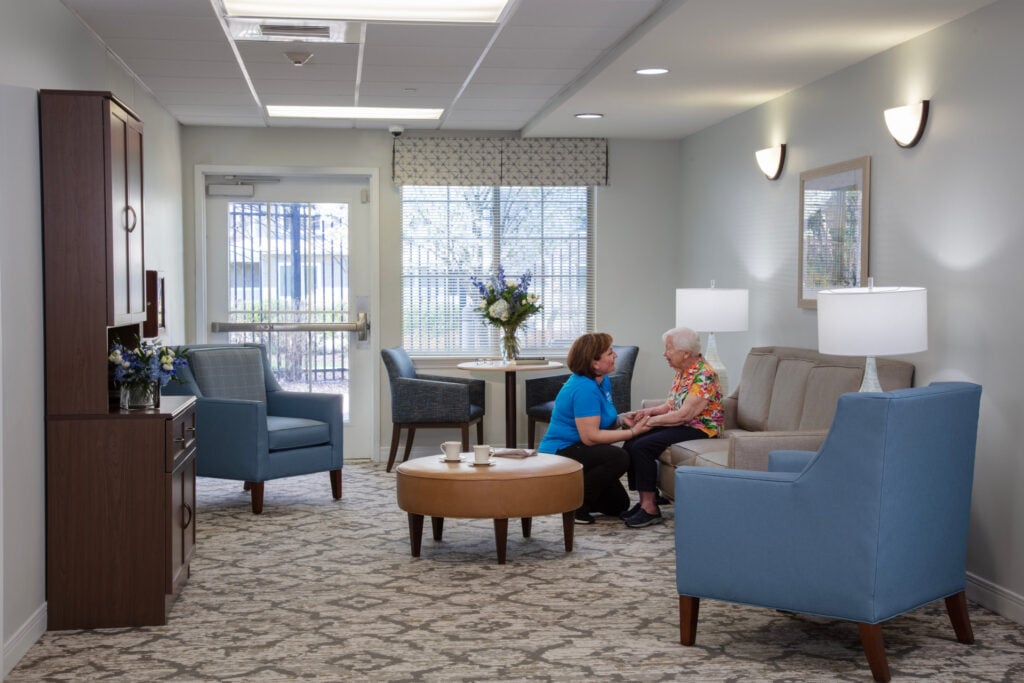 A caregiver in a blue polo shirt sits with an elderly resident on a sofa in a bright, warmly furnished common room lounge, engaged in friendly conversation.