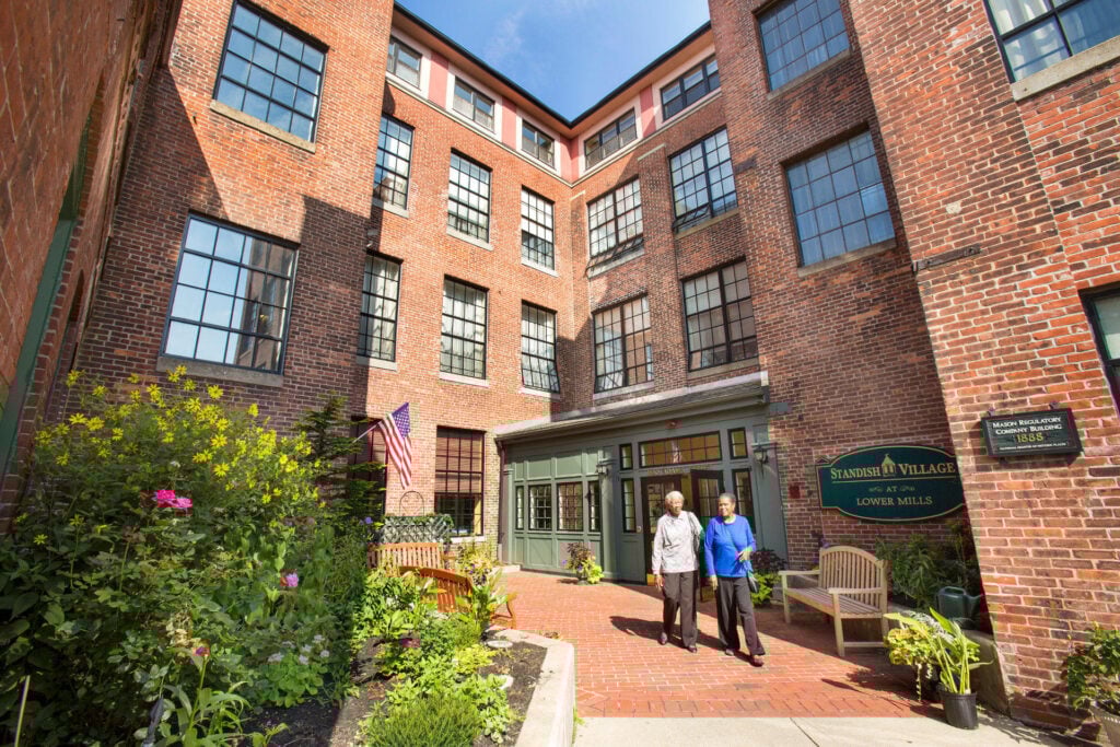 Two residents walk together along a brick courtyard pathway outside the entrance of Standish Village at Lower Mills, a red brick building with lush garden plantings and an American flag.