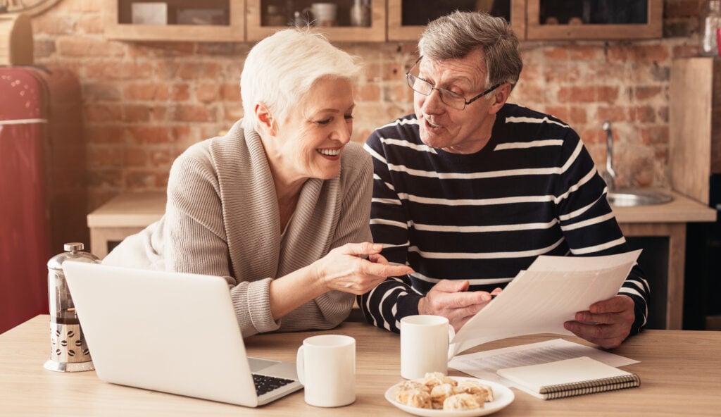 Happy Senior Couple Discussing Family Budget Together, Sitting In Kitchen With Laptop And Papers