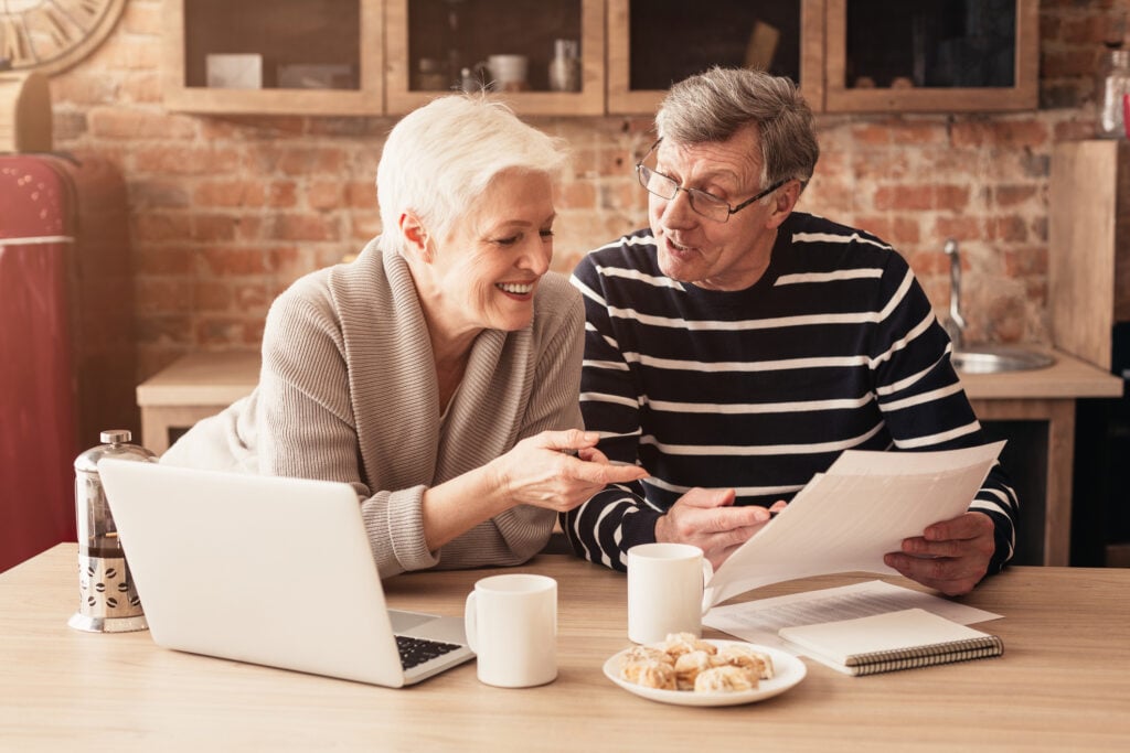Happy Senior Couple Discussing Family Budget Together, Sitting In Kitchen With Laptop And Papers