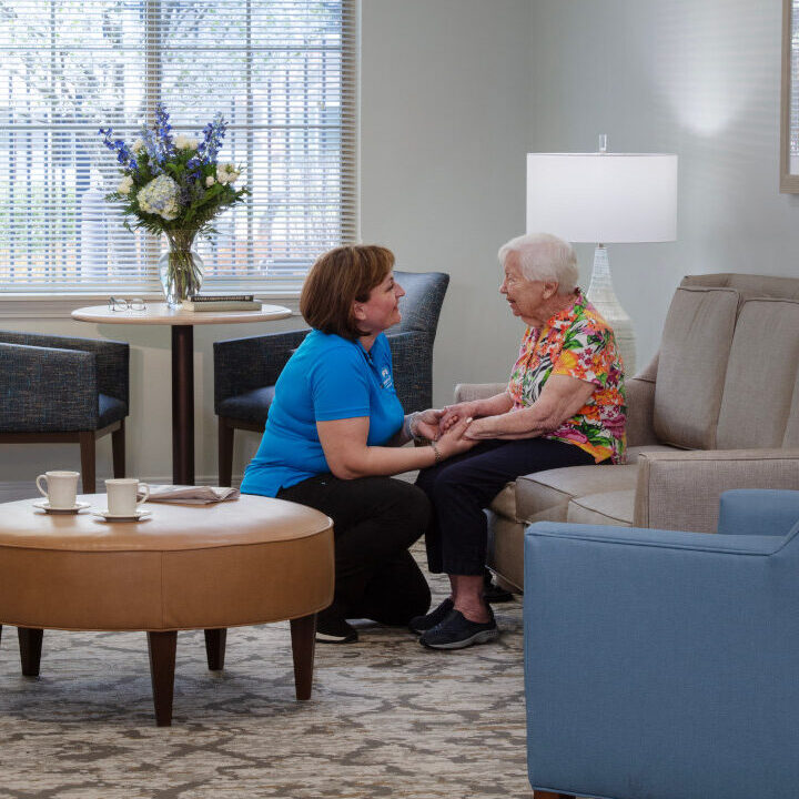 A caregiver in a blue polo shirt sits with an elderly resident on a sofa in a bright, warmly furnished common room lounge, engaged in friendly conversation.