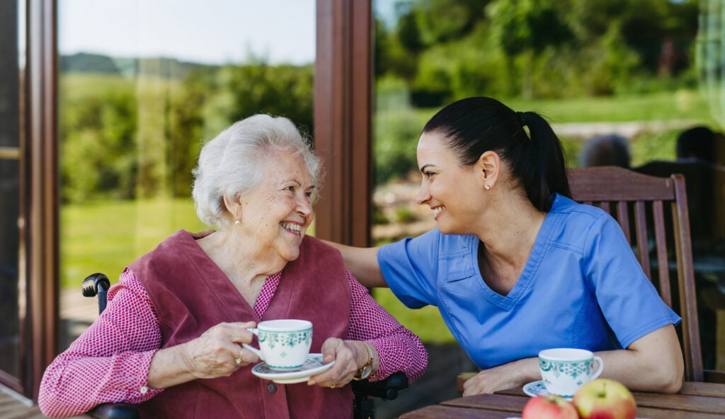 Caregiver spending time with elderly woman, drinking coffee, talking.